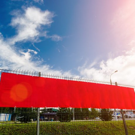 Red blank advertising banner hanging on a fence against the blue sky on a sunny day.